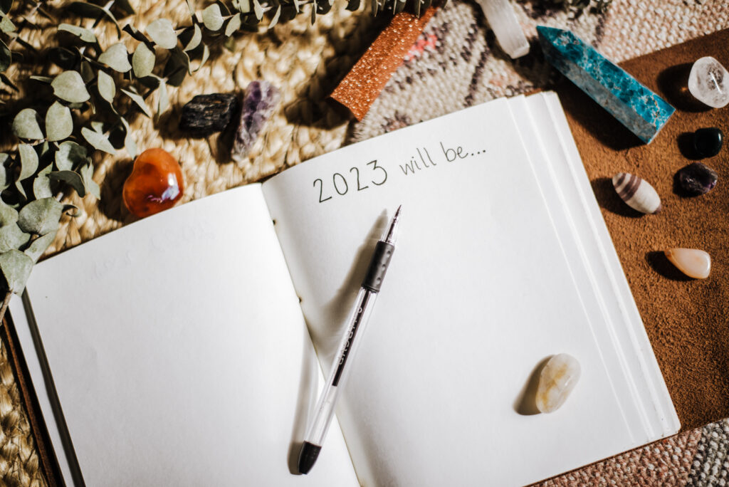 Open journal, mostly blank except for the words "2023 will be..." Black pen resting on top of journal, on a table surrounded by crystals